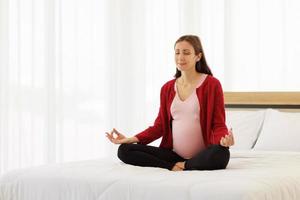 schwangere frau sitzt in ihrem schlafzimmer im bett und meditiert allein in frieden und glück. die neue Mutter ruht sich aus, indem sie sich hinsetzt, um sich zu entspannen. foto