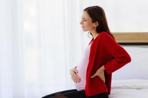 schwangere frau sitzt im schlafzimmer auf dem bett, hält ihren rücken und zeigt schmerzen oder schmerzenden rücken. foto