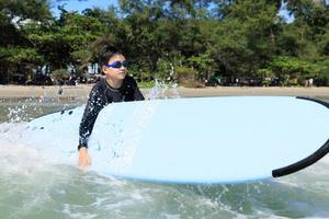 kleiner Junge, Student im Surfen, hält Softboard und versucht, es zurück ins Meer zu bringen, um zu üben, während er gegen Wellen und Spritzwasser spielt. foto