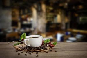 Heiße Kaffeetasse mit frischen organischen roten Kaffeebohnen und Kaffeeröstungen auf dem Holztisch im Hintergrund des Cafés