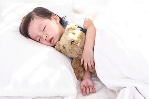 Kleines asiatisches Mädchen, das in ihrem Bett schläft. umarmt einen Teddybären foto