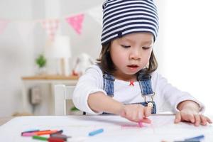 Kleines asiatisches Mädchen, das am Tisch im Zimmer sitzt, Vorschulmädchen, das an sonnigen Tagen mit bunten Stiften auf Papier zeichnet, Kindergarten oder foto