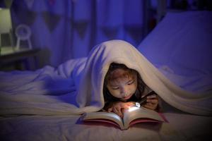 Kind asiatisches Mädchen, das nachts vor dem Schlafen ein Buch mit Taschenlampe unter der Decke auf dem Bett in einem dunklen Schlafzimmer liest, komfortables Konzept für Kinder zu Hause foto