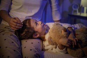 mutter streichelt den kopf eines süßen mädchens und schläft dann nachts auf dem mutterschoß auf dem bett in einem dunklen schlafzimmer, kind asiatisches mädchen umarmt teddybär, bequemes kinder zu hause konzept foto