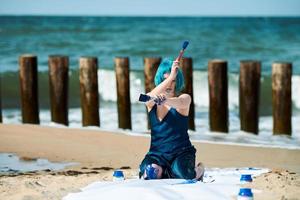 Künstlerische blauhaarige Performance-Künstlerin mit blauen Gouachefarben, die am Strand sitzen foto