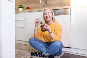 süße albino junge frau in modernen, stilvollen kleidern, die leckeren schokoladenaufstrich mit süßem lächeln in der küche genießen. junge Frau, die Schokolade aus einem Glas isst, während sie auf dem Küchenboden sitzt. foto