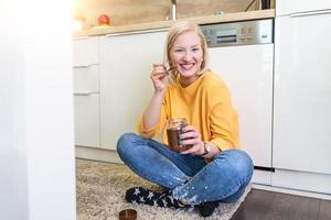 süße albino junge frau in modernen, stilvollen kleidern, die leckeren schokoladenaufstrich mit süßem lächeln in der küche genießen. junge Frau, die Schokolade aus einem Glas isst, während sie auf dem Küchenboden sitzt. foto