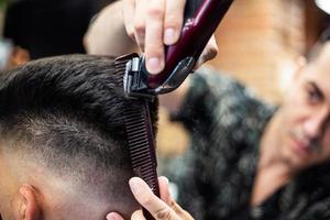 Rückansicht des jungen Mannes, der einen modernen Haarschnitt erhält. Mann wird mit professioneller elektrischer Haarschneidemaschine im Friseursalon getrimmt. männliches schönheitsbehandlungskonzept. kerl, der im friseursalon einen neuen haarschnitt bekommt foto