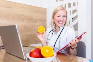 lächelnde ernährungsberaterin in ihrem büro zeigt sie gesundes gemüse und obst, gesundheits- und diätkonzept. weibliche ernährungsberaterin mit früchten, die an ihrem schreibtisch arbeitet. foto