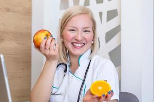 lächelnde ernährungsberaterin in ihrem büro zeigt sie gesundes gemüse und obst, gesundheits- und diätkonzept. weibliche ernährungsberaterin mit früchten, die an ihrem schreibtisch arbeitet. foto