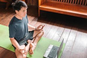 junger asiatischer mann coach internet video online training yoga lehrer moderner laptop bildschirm meditieren sukhasana haltung entspannen atmen leicht sitzen pose fitnessstudio gesunder lebensstil zu hause konzept. foto