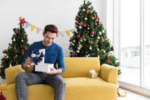 lächelnder hübscher junger mann, der zu hause weiße weihnachtsgeschenkbox öffnet foto