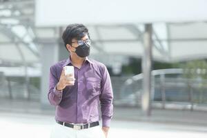 schwitzender asiatischer geschäftsmann mit gesichtsmaske mit handy während der bürozeiten als teil der neuen normalität des geschäfts foto