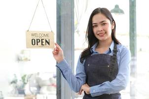 asiatische glückliche geschäftsfrau ist eine kellnerin in einer schürze, der besitzer des cafés steht mit einem offenen schild an der tür und wartet auf kunden. kleines Geschäftskonzept, Cafés und Restaurants foto