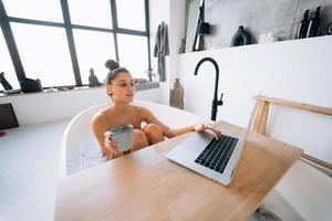 junge Frau, die am Laptop arbeitet, während sie eine Badewanne nimmt foto
