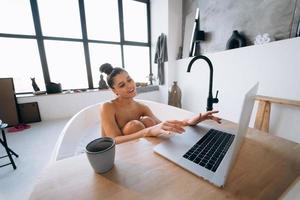 junge Frau, die am Laptop arbeitet, während sie eine Badewanne nimmt foto