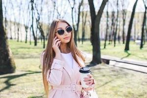 modische Frau mit Telefon und Kaffee in der Stadt. Modefrau in einer Sonnenbrille und einer rosa Jacke im Freien foto