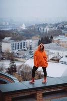 Mädchen in einer orangefarbenen Jacke posiert auf dem Dach eines Gebäudes im Stadtzentrum foto