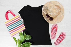 T-Shirt schwarz und Hausschuhe. t-shirt-modell flach lag mit sommeraccessoires. hut, tasche, rote flipflops und sonnenbrille auf holzbodenhintergrund. Platz kopieren. Vorlage leere Leinwand. Vorderansicht von oben. foto