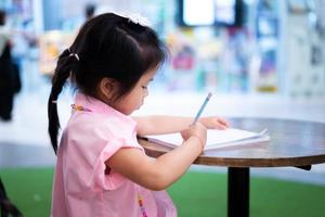 süßes asiatisches mädchen schreibt aufmerksam mit bleistift, kind übt kalligrafie, kinder machen hausaufgaben, die nach der schule vom lehrer zugewiesen werden, kind hält blauen bleistift mit ihrer rechten hand. foto