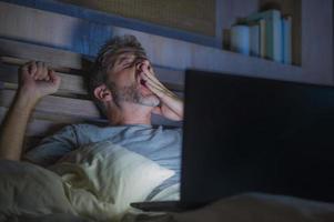 Attraktiver müder und gestresster Workaholic-Mann, der spät in der Nacht erschöpft auf dem Bett arbeitet und mit einem gähnenden Laptop-Computer beschäftigt ist, der sich schläfrig und überarbeitet fühlt foto