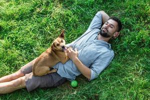 Attraktiver europäischer Mann ruht sich mit seinem süßen kleinen Hund an einem sonnigen Sommertag auf Gras aus. foto