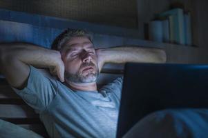Attraktiver müder und gestresster Workaholic-Mann, der spät in der Nacht erschöpft auf dem Bett arbeitet und mit einem Laptop beschäftigt ist, der sich schläfrig und überarbeitet fühlt foto