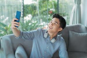 Selfie. Porträt eines jungen schönen Mannes mit blauem Hemd und blauen Kopfhörern macht ein Foto von sich selbst auf dem Handy