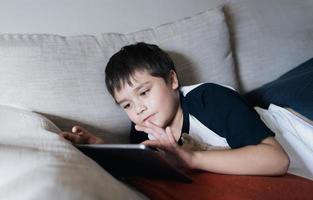 Schuljunge, der ein Tablet verwendet, um online im Internet zu lernen, ein Kind, das ein Spiel spielt oder Cartoons auf einem digitalen Pad ansieht, ein glückliches Kind, das sich zu Hause entspannt, Kinder mit Technologie und einem Konzept für soziale Netzwerke foto