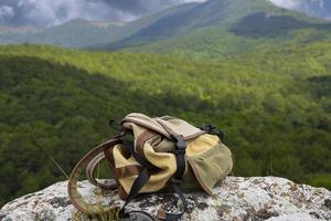 Ein braungrüner Rucksack liegt auf einem großen grauen Stein auf einem Felsen vor dem Hintergrund grüner Berge. foto