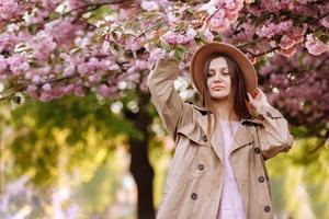 Porträt eines jungen schönen modischen Mädchens mit Hut, das an einem sonnigen Tag in der Nähe eines blühenden Baumes mit rosa Blüten posiert foto