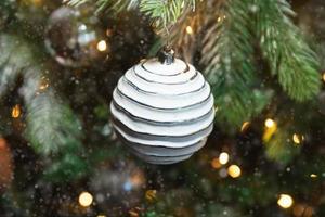 klassisches weihnachtsneujahr geschmückter neujahrsbaum mit weißen ornamentdekorationen spielzeug und ball, schnee und defokussierten girlandenlichtern. modernes klassisches Urlaubsdesign. Heiligabend zu Hause.