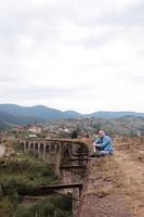 stilvoller junger mann auf der eisenbahnbrücke in worokhta, karpaten vor dem hintergrund einer berglandschaft. touristen sitzen in den bergen in der nähe der eisenbahn foto