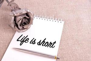 Das Leben ist kurz, der Text ist in ein Notizbuch geschrieben, das auf einem Leinentuch und einer eisernen Rosenblüte liegt. foto
