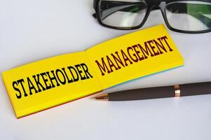 Stakeholder-Management-Text auf gelbem Notizblock mit Brille und Stift auf weißem Einbandhintergrund. Geschäftsbeziehungskonzept foto