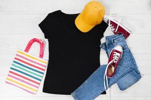 T-Shirt schwarz und Turnschuhe. t-shirt-modell flach lag mit sommeraccessoires. Baseballmütze, Tasche, gelbe Flip-Flops auf Holzbodenhintergrund. Platz kopieren. Vorlage leere Leinwand. Vorderansicht von oben. foto