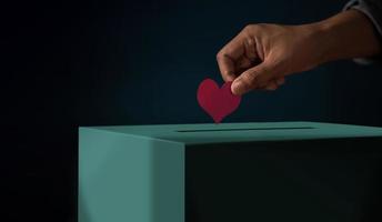 Spendenkonzept. Hand, die ein rotes Herzpapier in eine Spendenbox legt. Metapher Foto. dunkler Ton foto