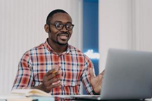 Lächelnder, verwirrter afroamerikanischer Mann, der E-Mails mit Nachrichten liest, am Laptop arbeitet und die Hände ausbreitet foto