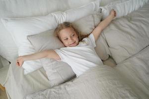 glückliches kleines Mädchen, das sich ausdehnt und nach dem Aufwachen im weichen Bett liegt. Gesunder Kinderschlaf foto