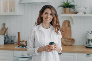 Lächelnde hispanische Frau mit Pappbecher Kaffee, die in einer gemütlichen, modernen Küche steht und in die Kamera blickt foto