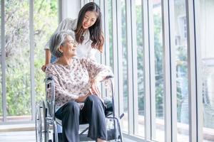 schöne asiatische Mädchen helfen und kümmern sich um ältere ältere Frauen, die im Wohnzimmer im Haus im Rollstuhl sitzen.
