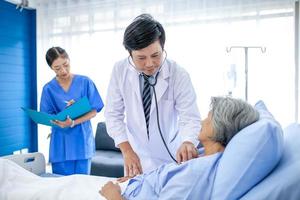 arzt, der die station besucht, hat eine patientin und überprüft sie mit einem stethoskop auf dem bett. Patientin liegt auf Krankenhausbett, während der Arzt ihre Untersuchung durchführt. foto