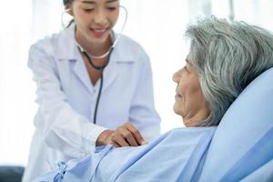 arzt, der die station besucht, hat eine patientin und überprüft sie mit einem stethoskop auf dem bett. Patientin liegt auf Krankenhausbett, während der Arzt ihre Untersuchung durchführt. foto