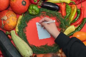 Gemüse wird um ein Blatt Papier und einen Bleistift gelegt. leerer Platz für Text. weibliche Hand, die ein Rezept auf einem leeren Rohling auf rotem Hintergrund schreibt. foto