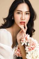 asiatische frau mit einem schönen gesicht und perfekt sauberer frischer haut mit blume. süßes weibliches Model mit natürlichem Make-up und funkelnden Augen auf beige isoliertem Hintergrund. foto