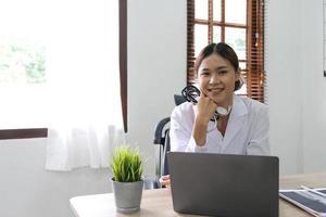 Lächelnde asiatische Ärztin mit Laptop-Computer in ihrem Büro. Freundlicher Arzt mit Tablet-Computer in der Klinik. foto