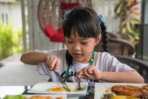 Kleines asiatisches Mädchen isst Spiegelei auf Teller am Tisch. foto