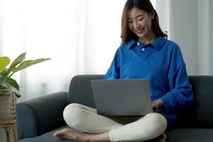 junge freiberufliche asiatische frau, die am laptop arbeitet und soziale medien überprüft, während sie auf dem sofa liegt, wenn sie sich im wohnzimmer zu hause entspannen. foto