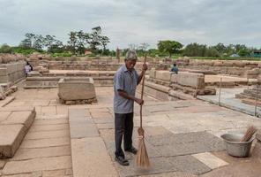 mamallapuram, tamil nadu, indien - september 2018 ein älterer indischer mann fegt den steinboden mit einem strohbesen der antiken ruinen des küstentempels im unesco-kulturerbe mahabalipuram. foto