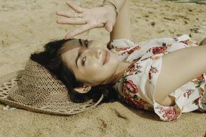Eine junge schöne asiatische Frau, die vom Licht geblendet wird, während sie auf dem Strandsand liegt foto